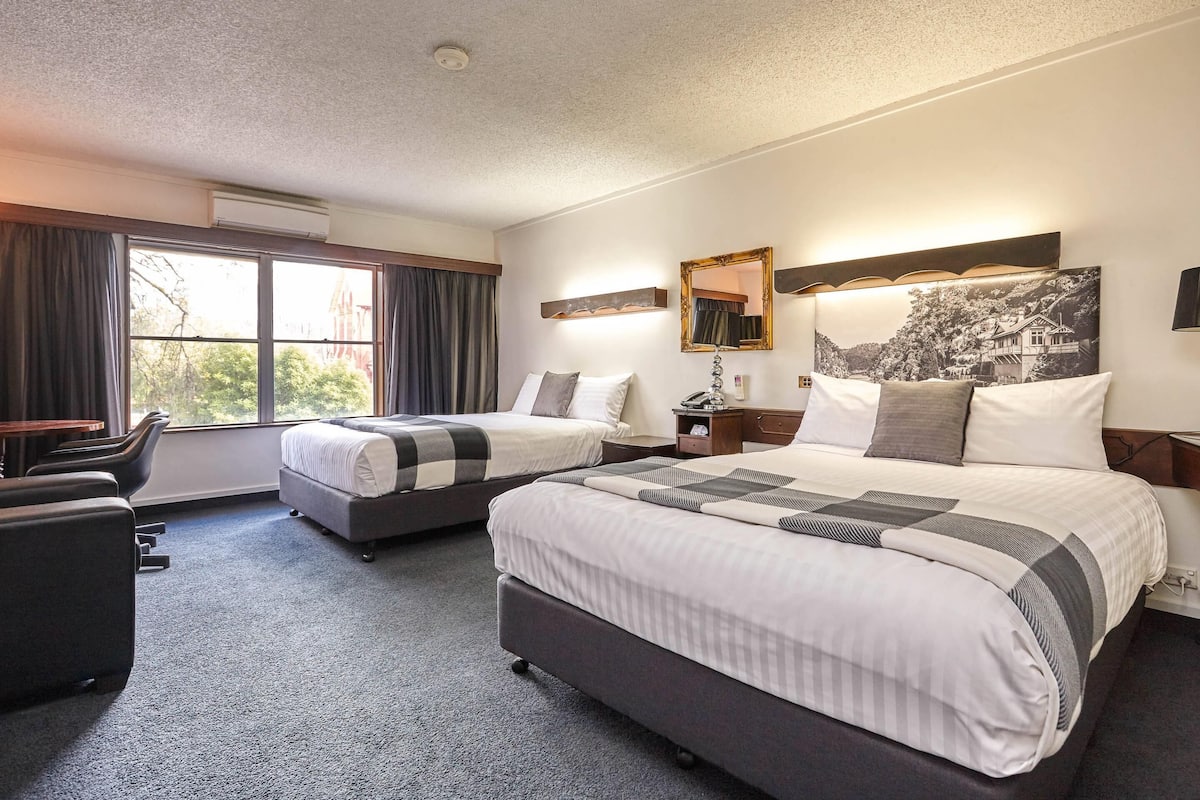 A hotel room with two beds, a desk, a chair, a mirror, and a large window.