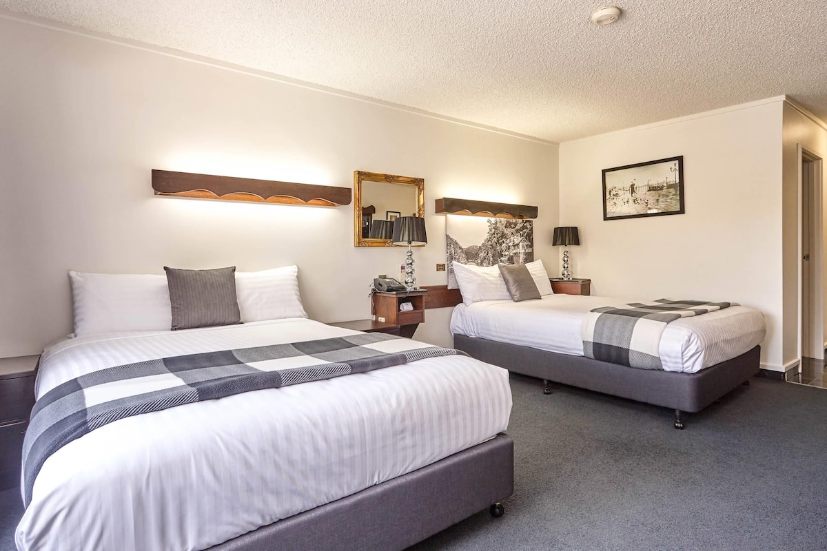 A hotel room with two beds, a nightstand with a lamp, a wall-mounted mirror, and a framed picture.