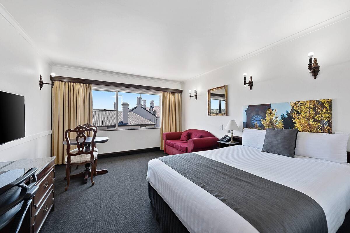 A hotel room with a large bed, a desk with drawers, a chair, a red sofa, a window with curtains, and a painting on the wall.