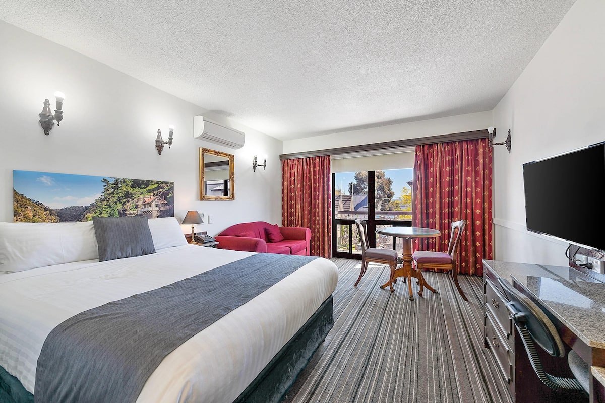 A hotel room with a large bed, a red armchair, a small table, a TV, and a balcony with red curtains.