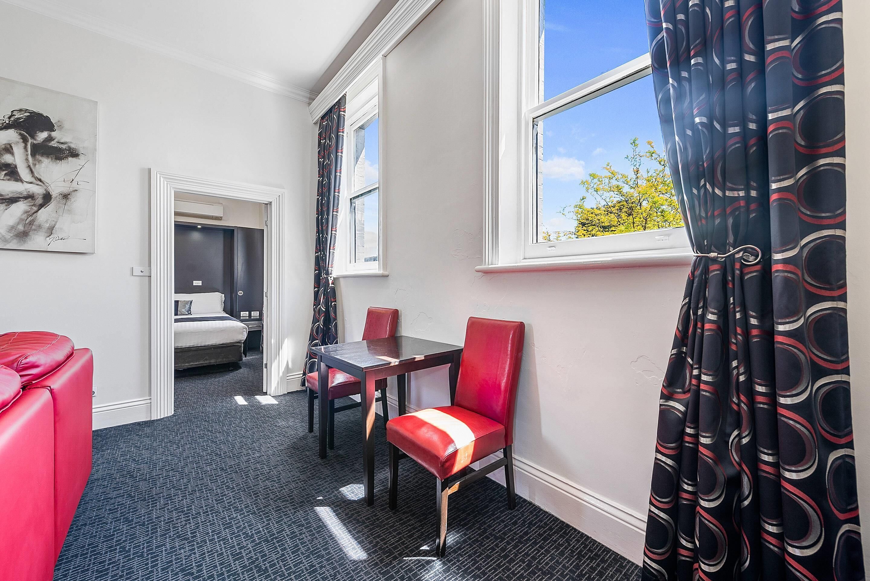 Une chambre d’hôtel avec une table à manger, des chaises rouges, un canapé rouge, un lit et une grande fenêtre avec des rideaux.