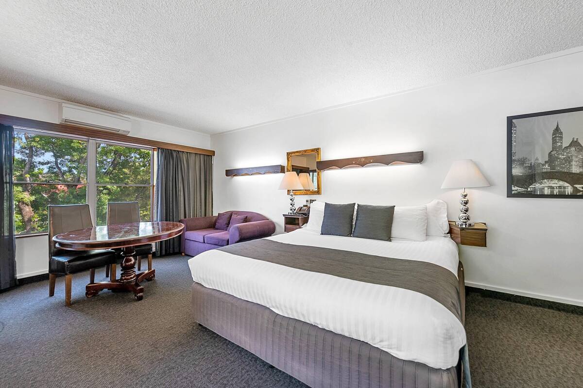 A hotel room with a large bed, a round table, a purple armchair, and a cityscape painting.