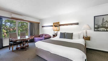 A hotel room with a large bed, a round table, a purple armchair, and a cityscape painting.