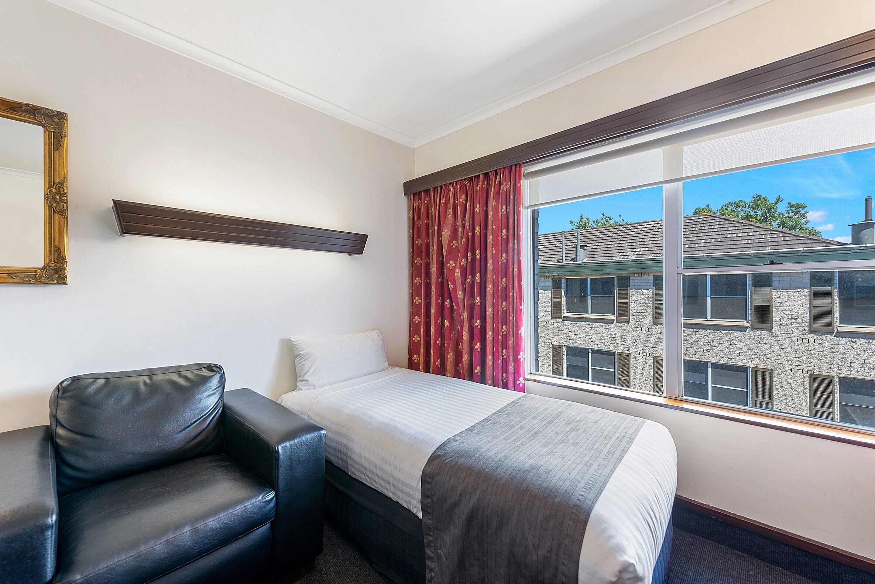 Une chambre d’hôtel avec un lit, une chaise, une fenêtre avec des rideaux, un miroir et une étagère.