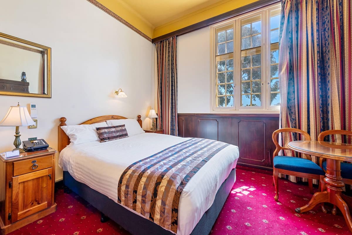 A neatly made bed with a patterned blanket, a bedside table with a lamp, a wooden nightstand, a chair, and a window with curtains.