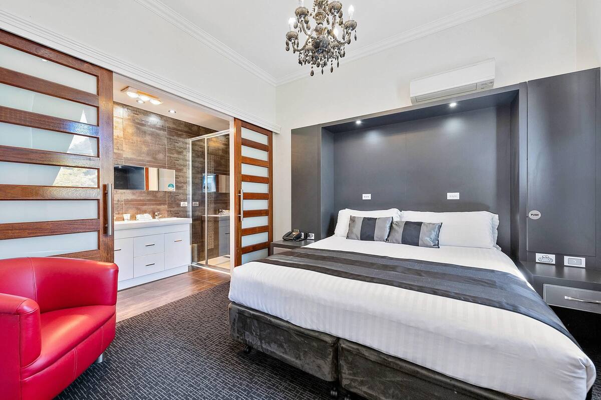 A modern hotel room with a large bed, a red chair, a glass partition, and a chandelier.