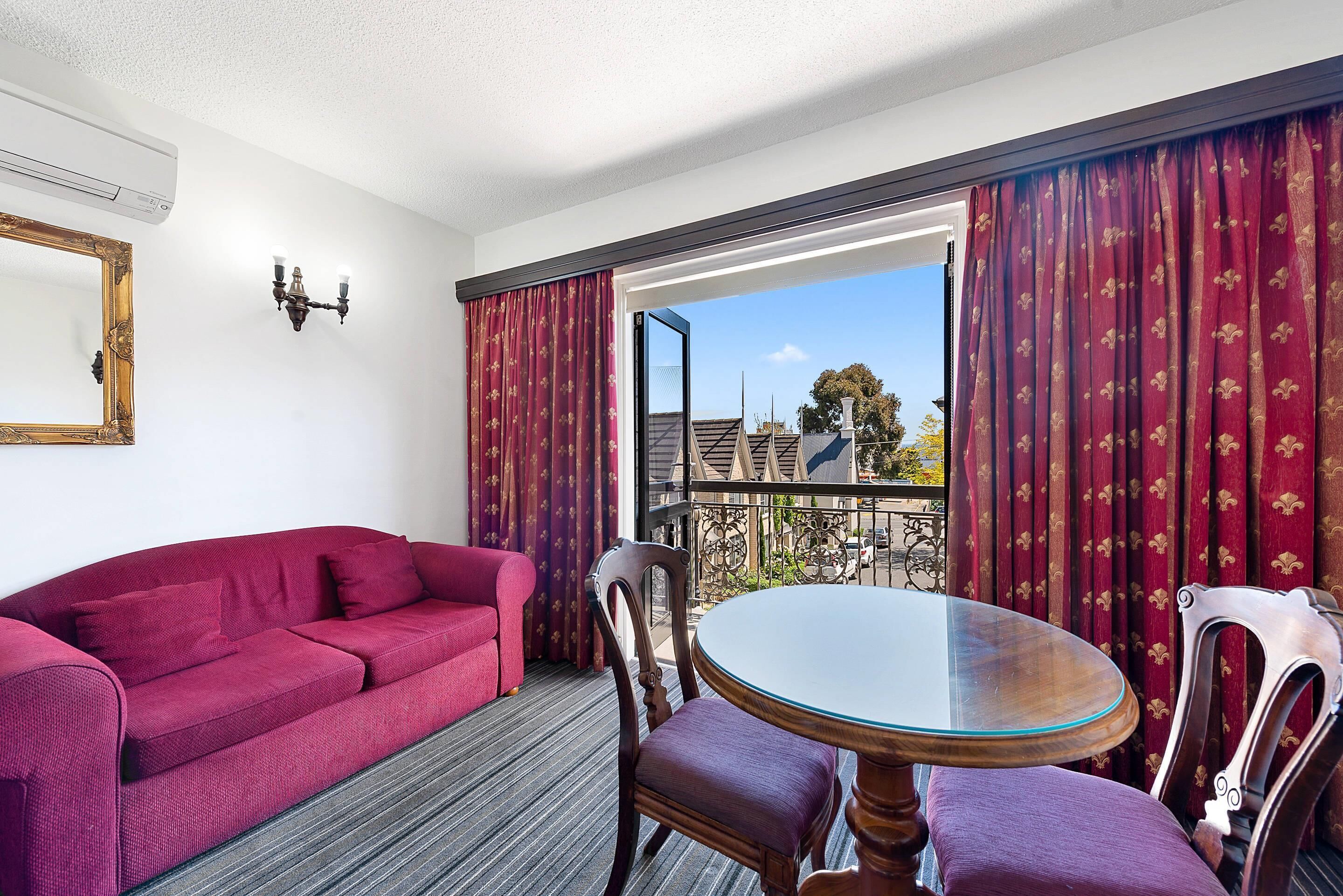 A room with a red sofa, a round table with chairs, a chandelier, and a large window with curtains.