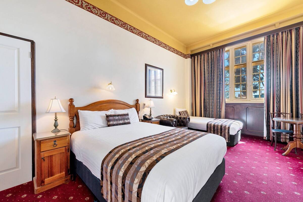 A hotel room with two beds, a wooden nightstand, a lamp, a framed picture on the wall, a window with curtains, and a red carpet.