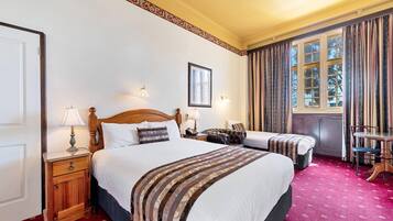 A hotel room with two beds, a wooden nightstand, a lamp, a framed picture on the wall, a window with curtains, and a red carpet.