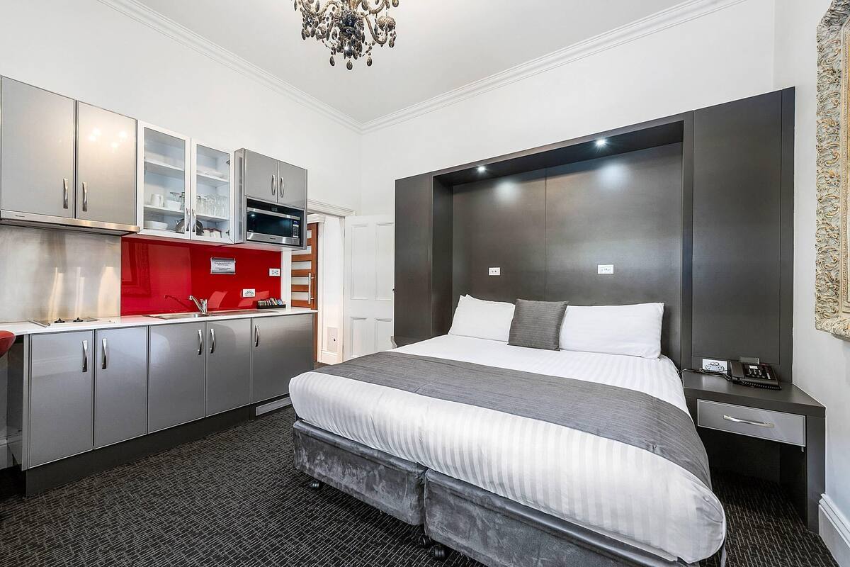 A modern hotel room with a large bed, a built-in desk, a kitchenette, and a chandelier.