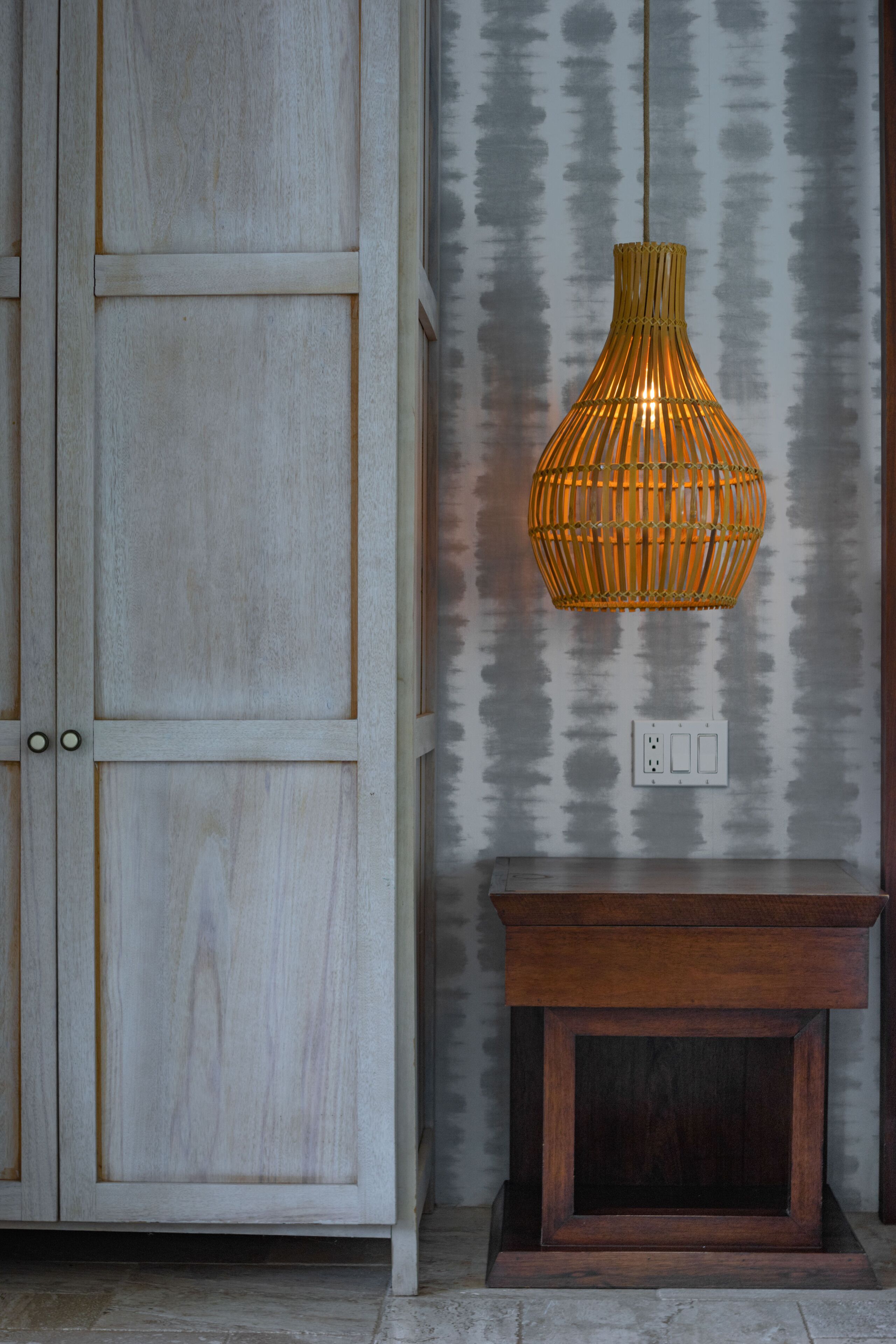 A wooden cabinet with a light finish, a small wooden table, and a hanging wicker lamp against a textured wall.