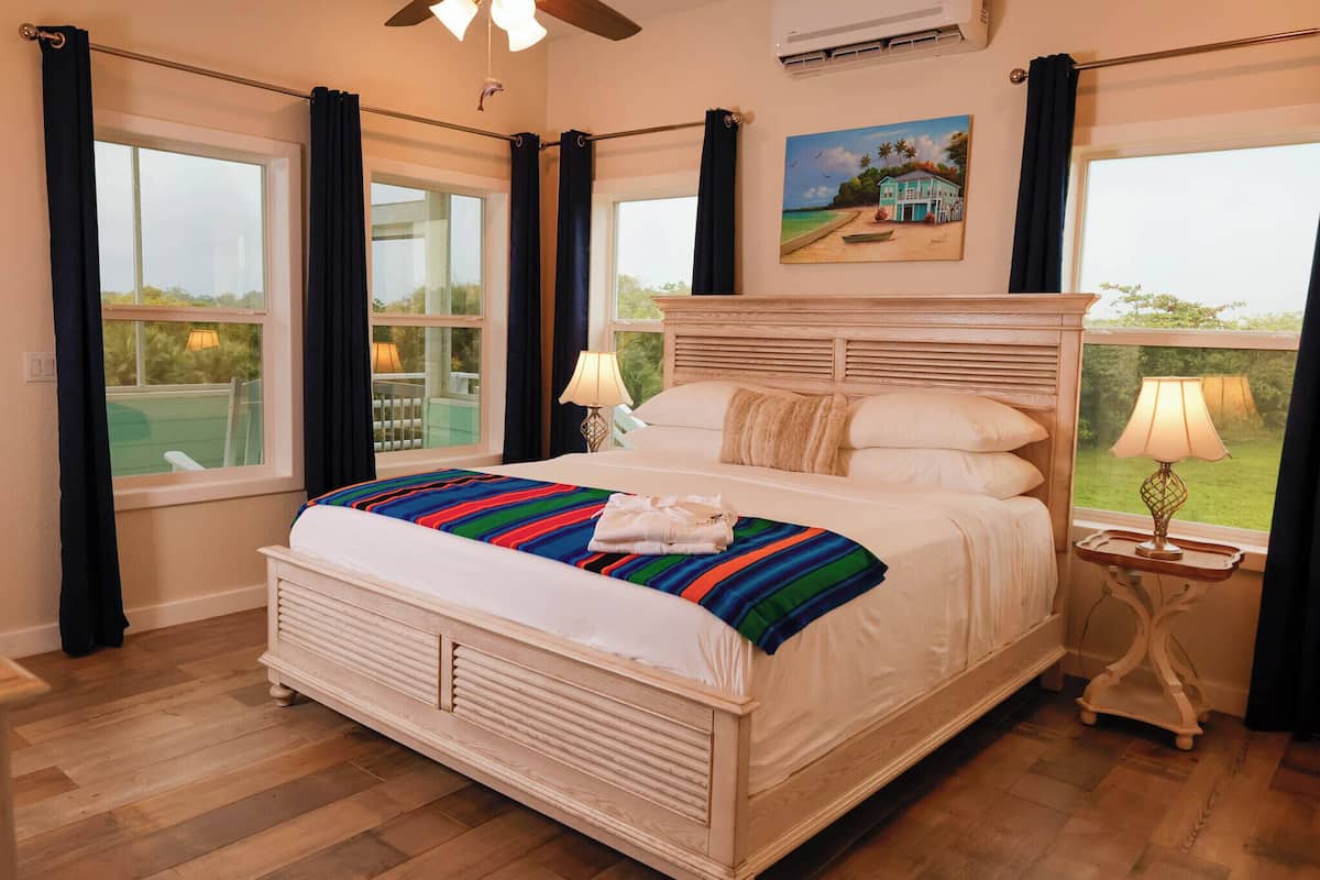 A bedroom with a large bed, two bedside lamps, a window with curtains, and a painting on the wall.