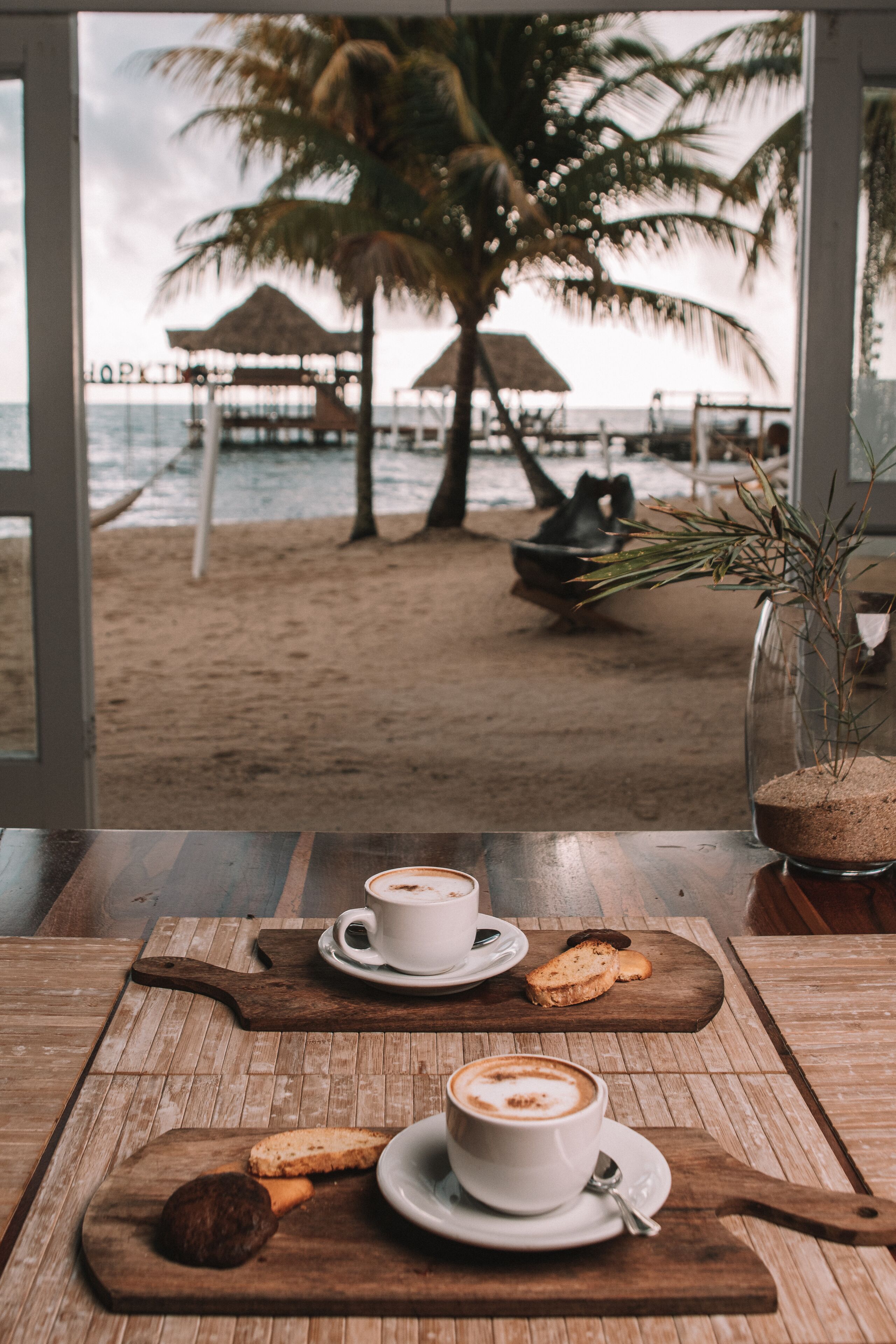 A table with a cup of coffee, a pastry, and a cookie on a wooden board, with a view of a beach and palm trees through a window.