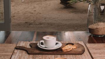 A table with a cup of coffee, a pastry, and a cookie on a wooden board, with a view of a beach and palm trees through a window.