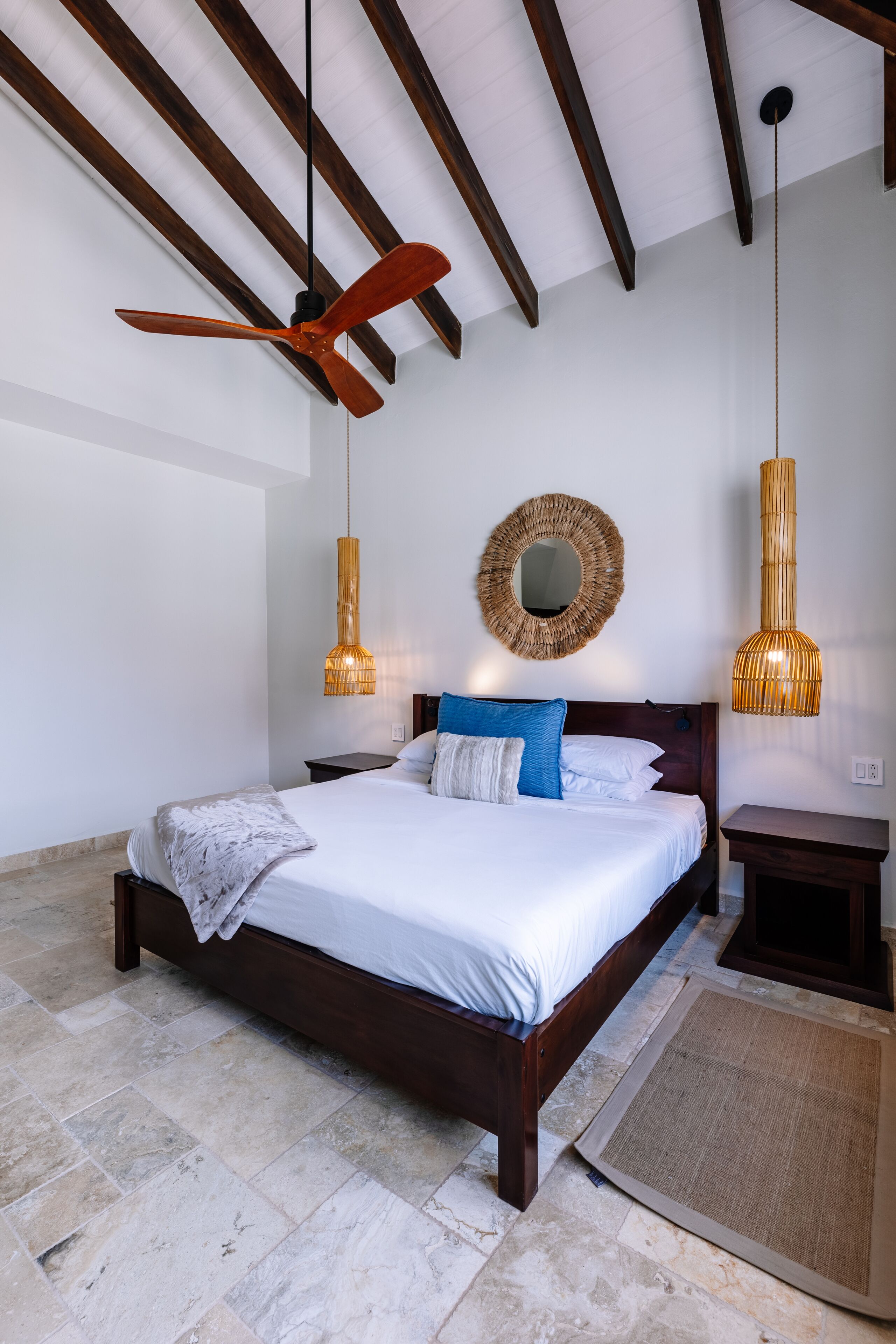 A bedroom with a wooden bed, a ceiling fan, a woven mirror, and two hanging lamps.
