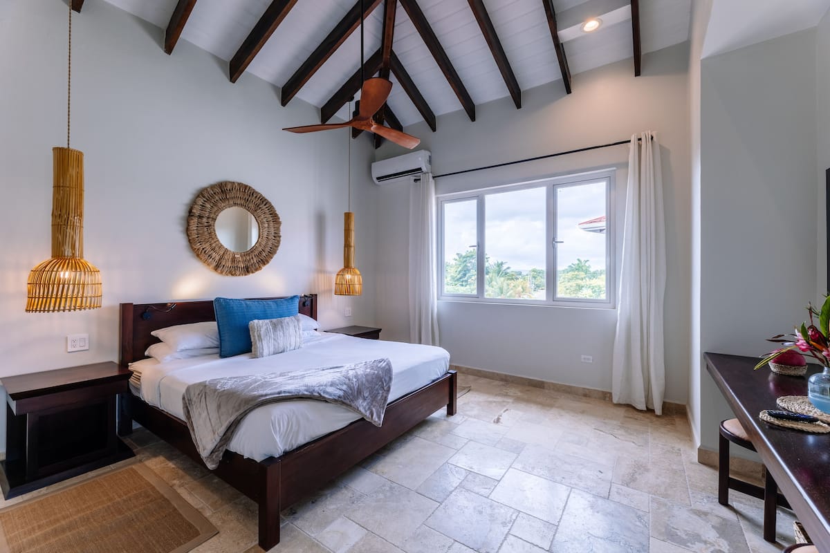A bedroom with a bed, bedside tables, a ceiling fan, a window with curtains, and a view of greenery outside.