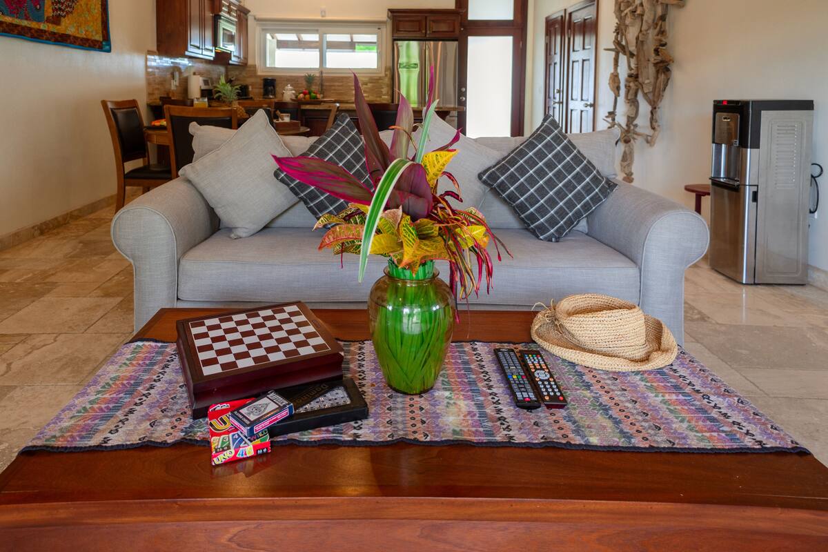 A living room with a sofa, a coffee table with a chess set, a vase with plants, and a remote control.