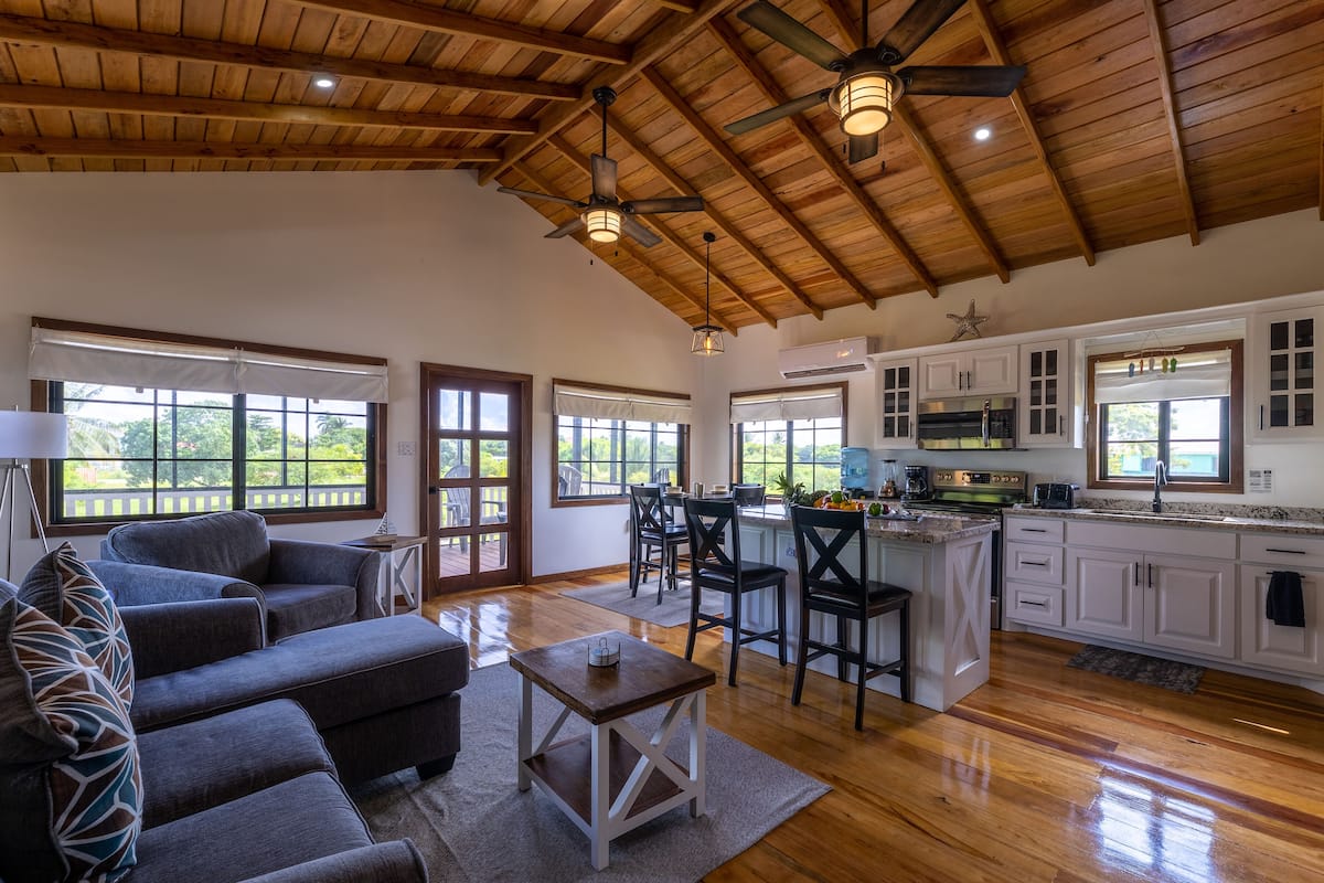 A spacious living area with a wooden ceiling, a kitchen with white cabinets, and a large window overlooking greenery.
