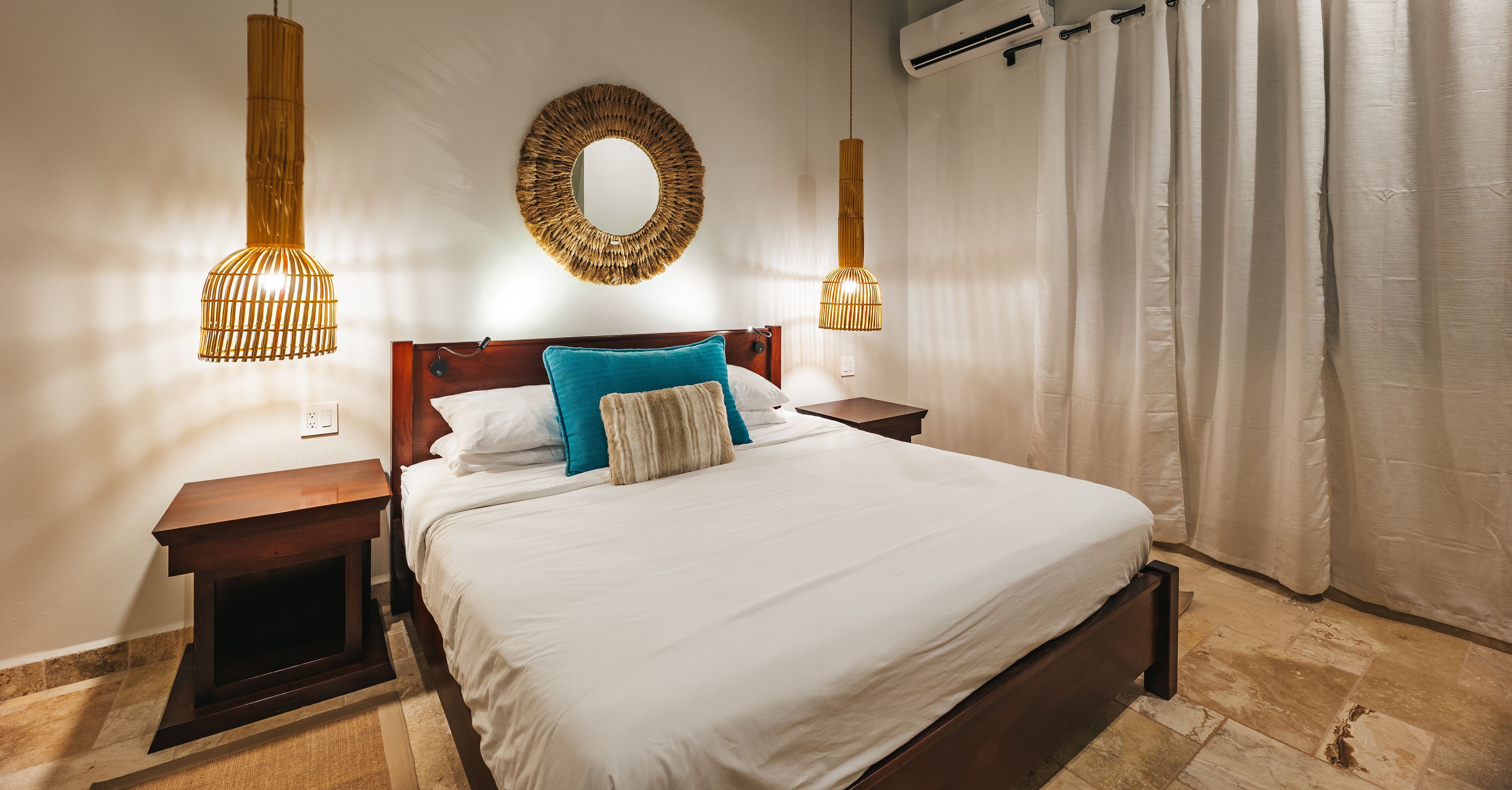 A neatly made bed with white bedding and a blue pillow, flanked by two bedside tables with lamps, in a room with a large mirror and an air conditioning unit on the wall.