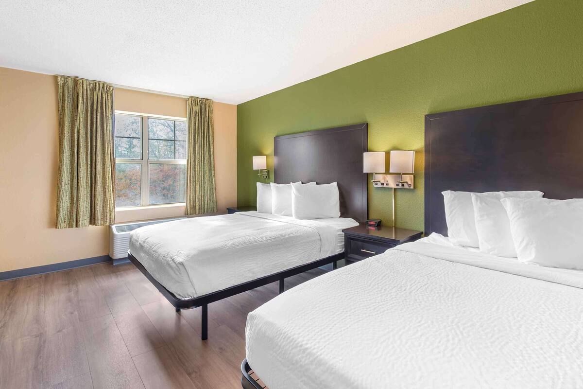 A hotel room with two beds, a large window with curtains, a headboard, bedside lamps, and a wall-mounted heater.