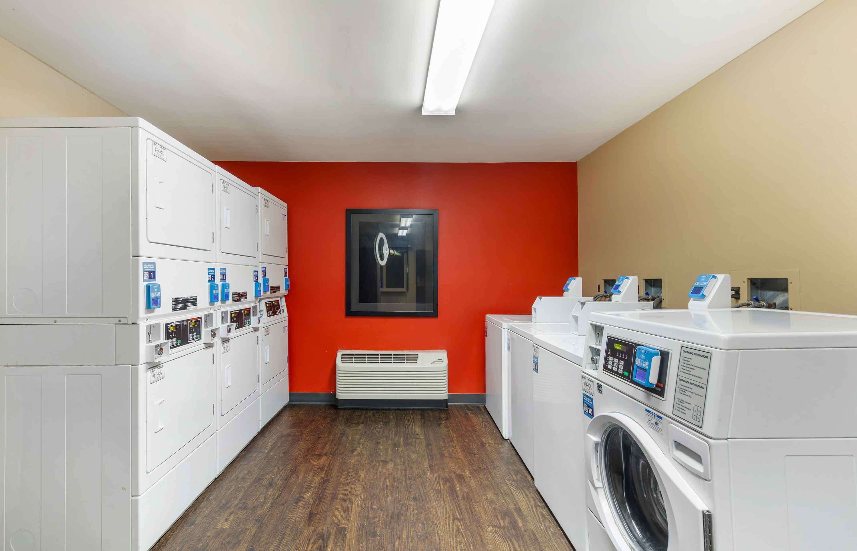 A laundry room with multiple washing machines and dryers, a red wall, and a ceiling light.