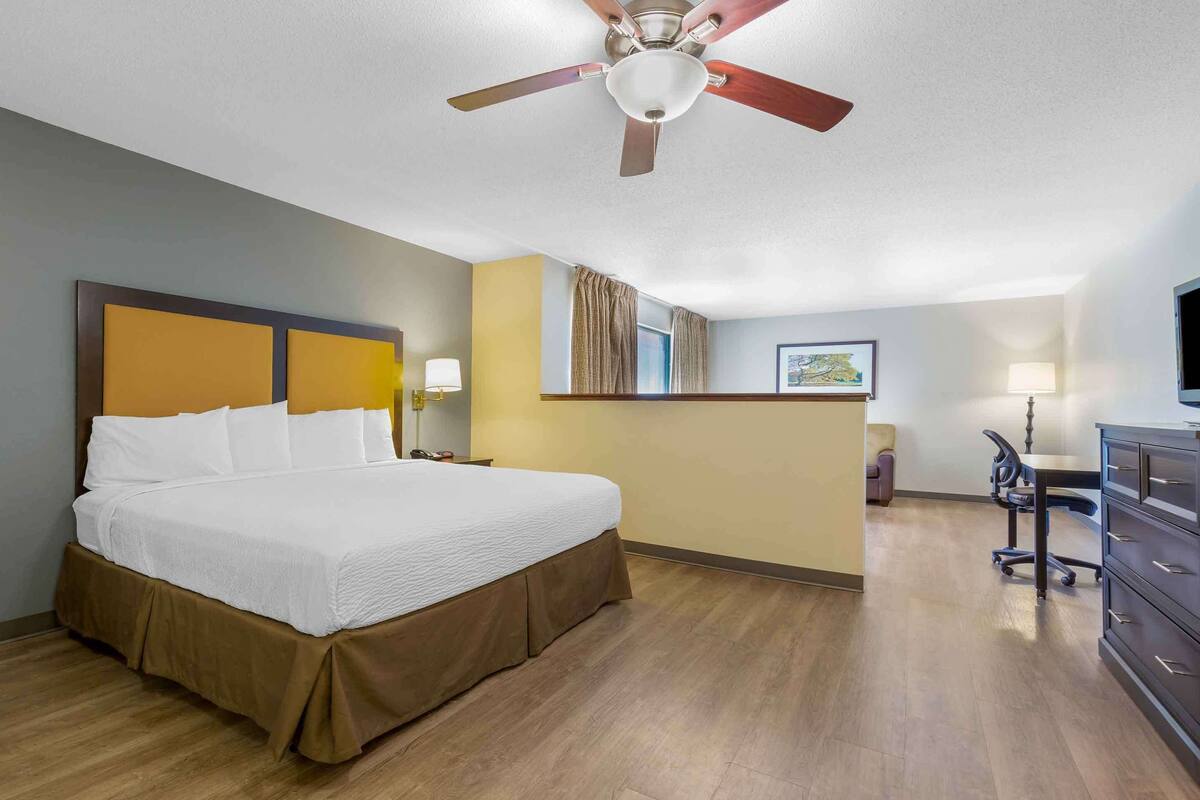 A hotel room with a large bed, a desk, a chair, a ceiling fan, and a television.