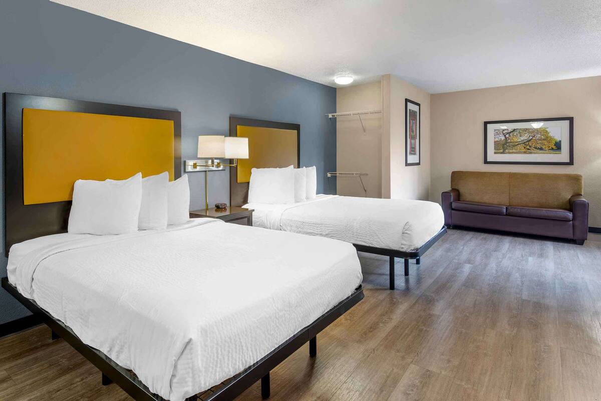 A hotel room with two beds, a sofa, a nightstand with a lamp, and a framed picture on the wall.