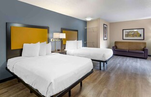 Habitación de hotel con dos camas, un sofá, una mesita de noche con lámpara y un cuadro en la pared.