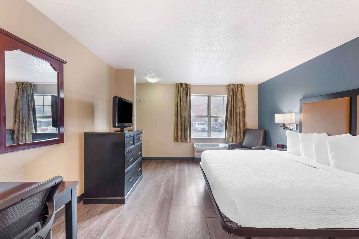 A hotel room with a large bed, a desk with a chair, a TV, a dresser, and a window with curtains.