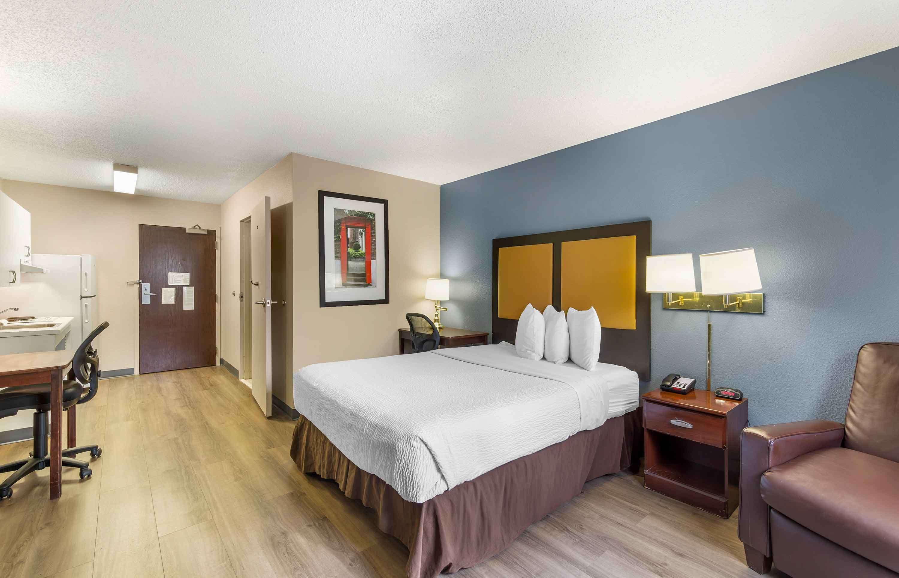 A hotel room with a bed, bedside table, lamp, chair, desk, and a door leading to another room.