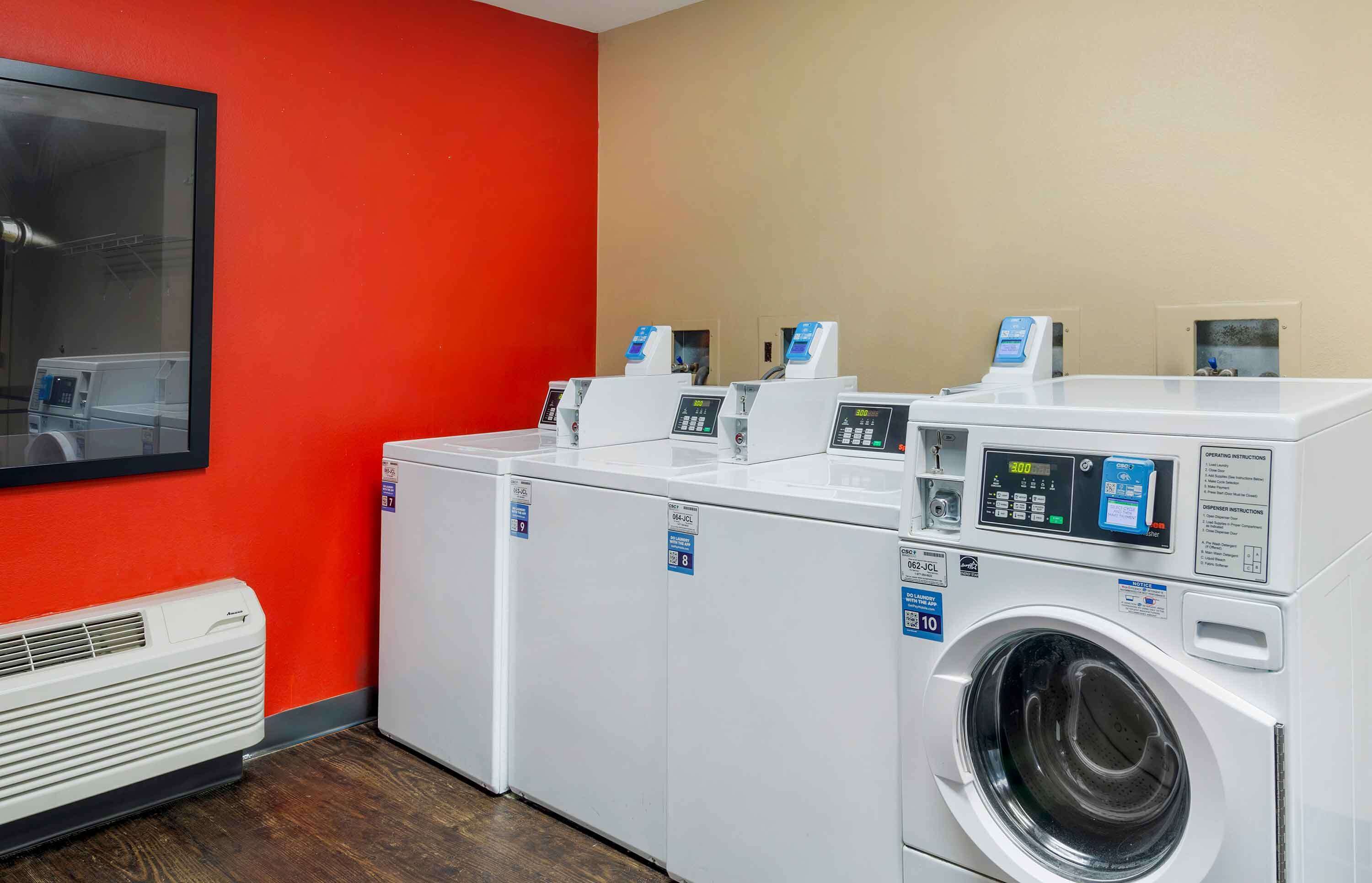 A laundry facility with multiple washing machines and dryers, a wall-mounted air conditioning unit, and a large window.
