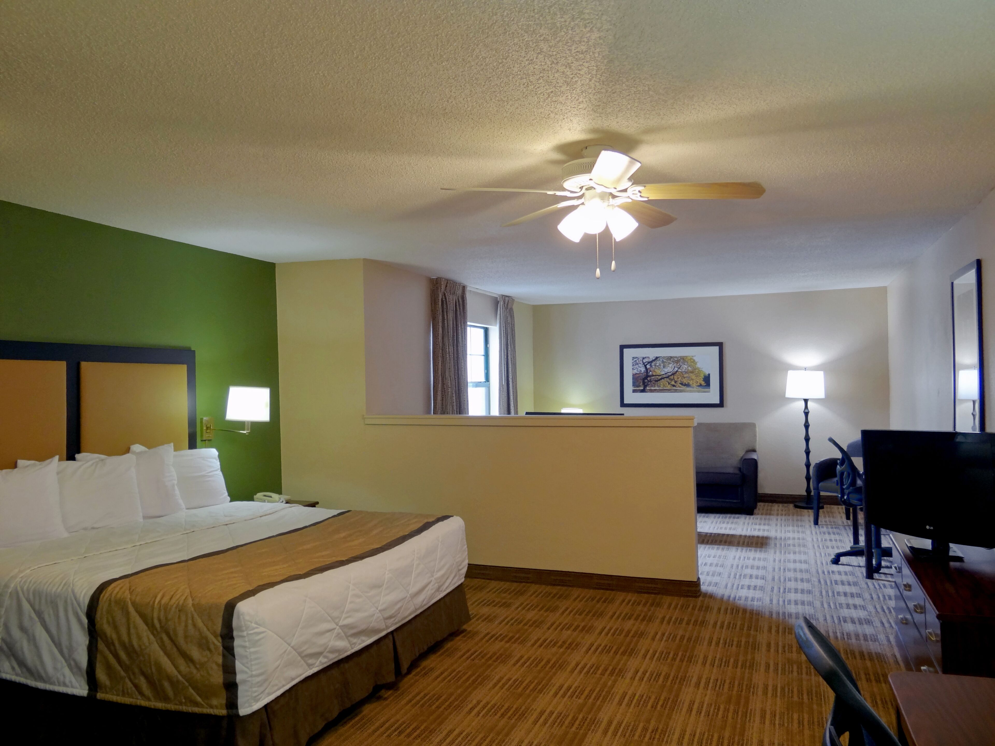 Una habitación de hotel con una cama, un escritorio, una silla, un televisor, un ventilador de techo, una ventana con cortinas y un cuadro en la pared.
