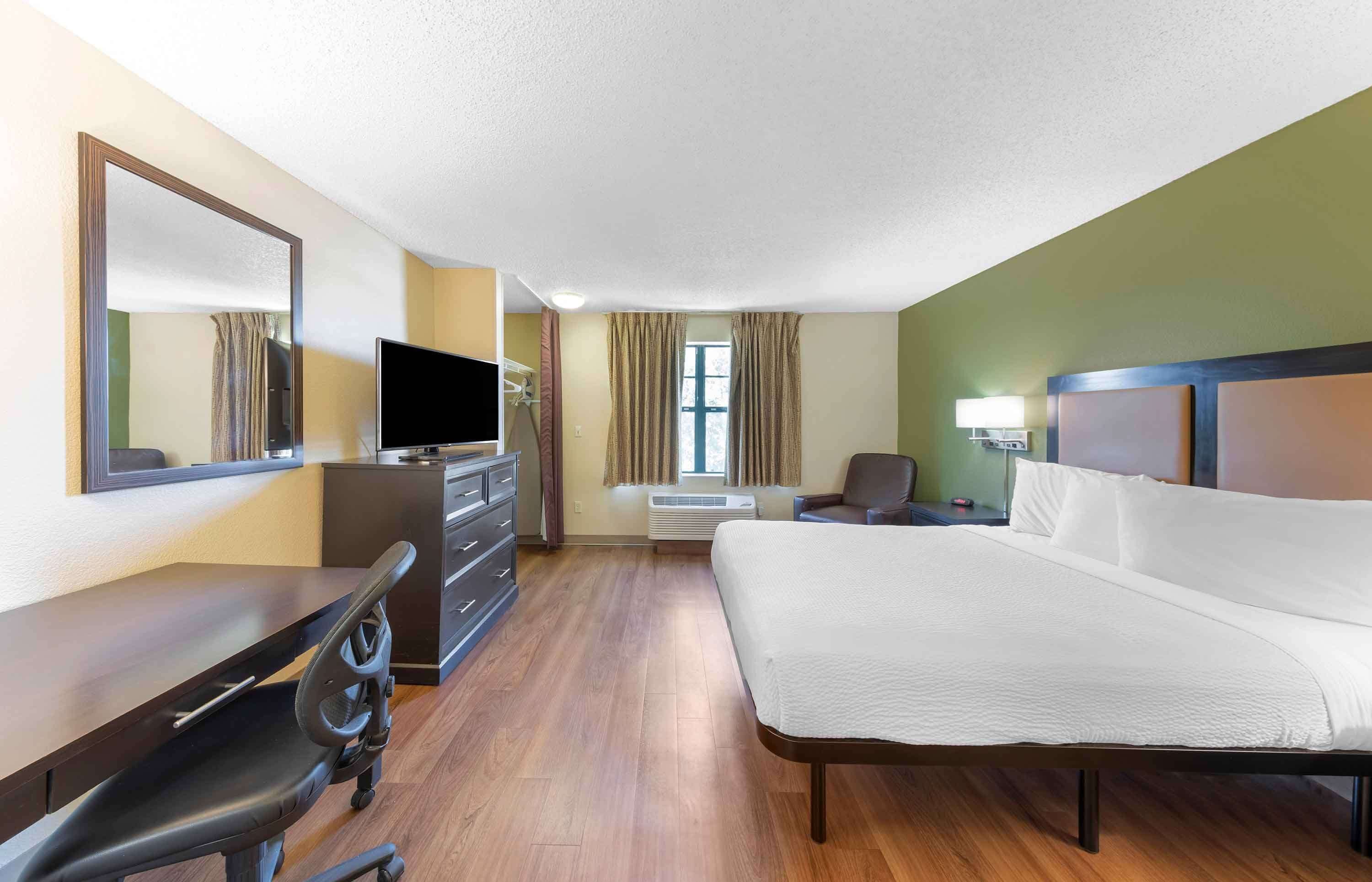 Ein Hotelzimmer mit einem Bett, einem Schreibtisch mit Stuhl, einem Fernseher, einer Kommode und einem Spiegel.