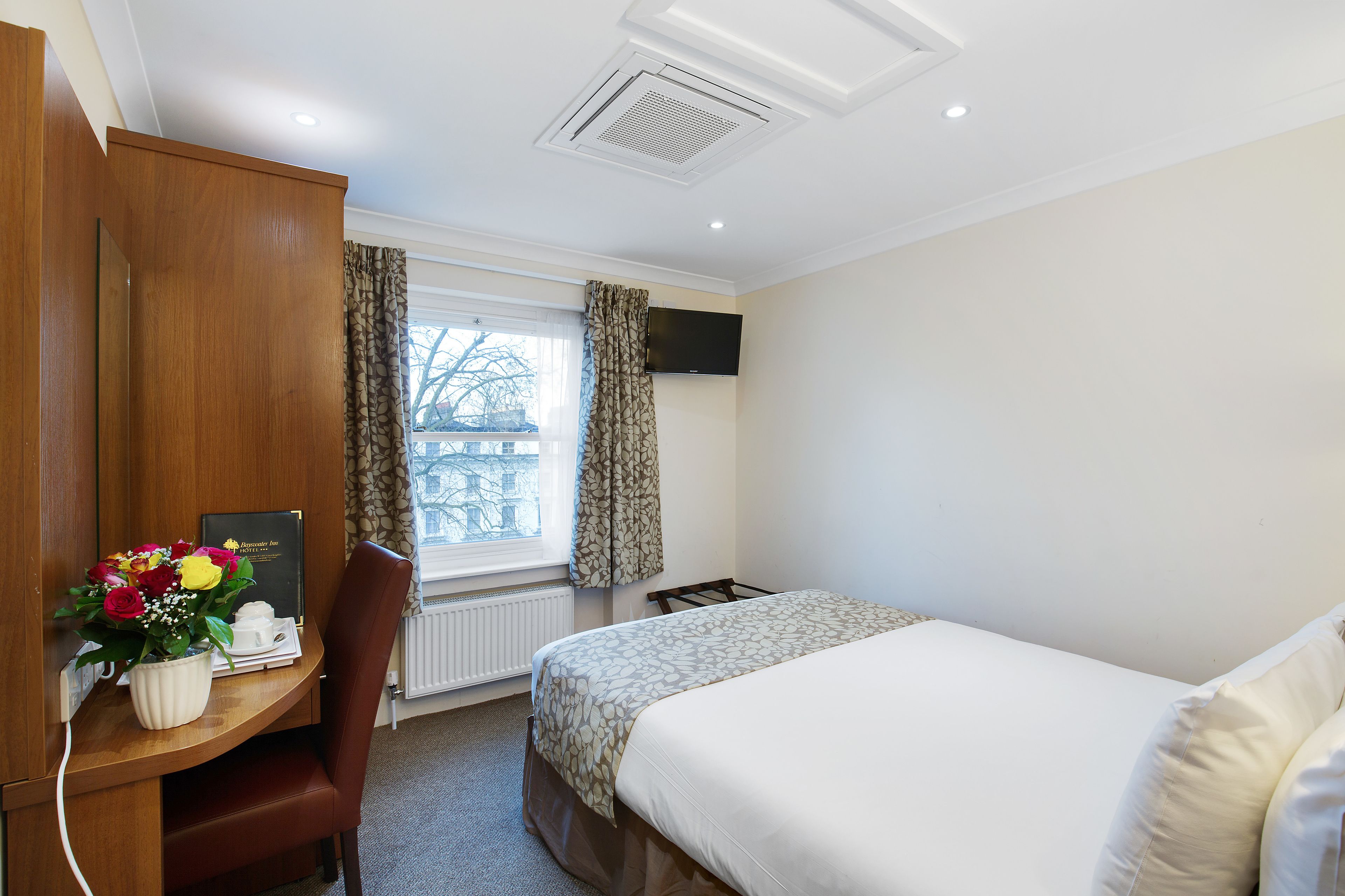 A hotel room with a bed, a desk with a chair, a TV mounted on the wall, and a window with curtains.
