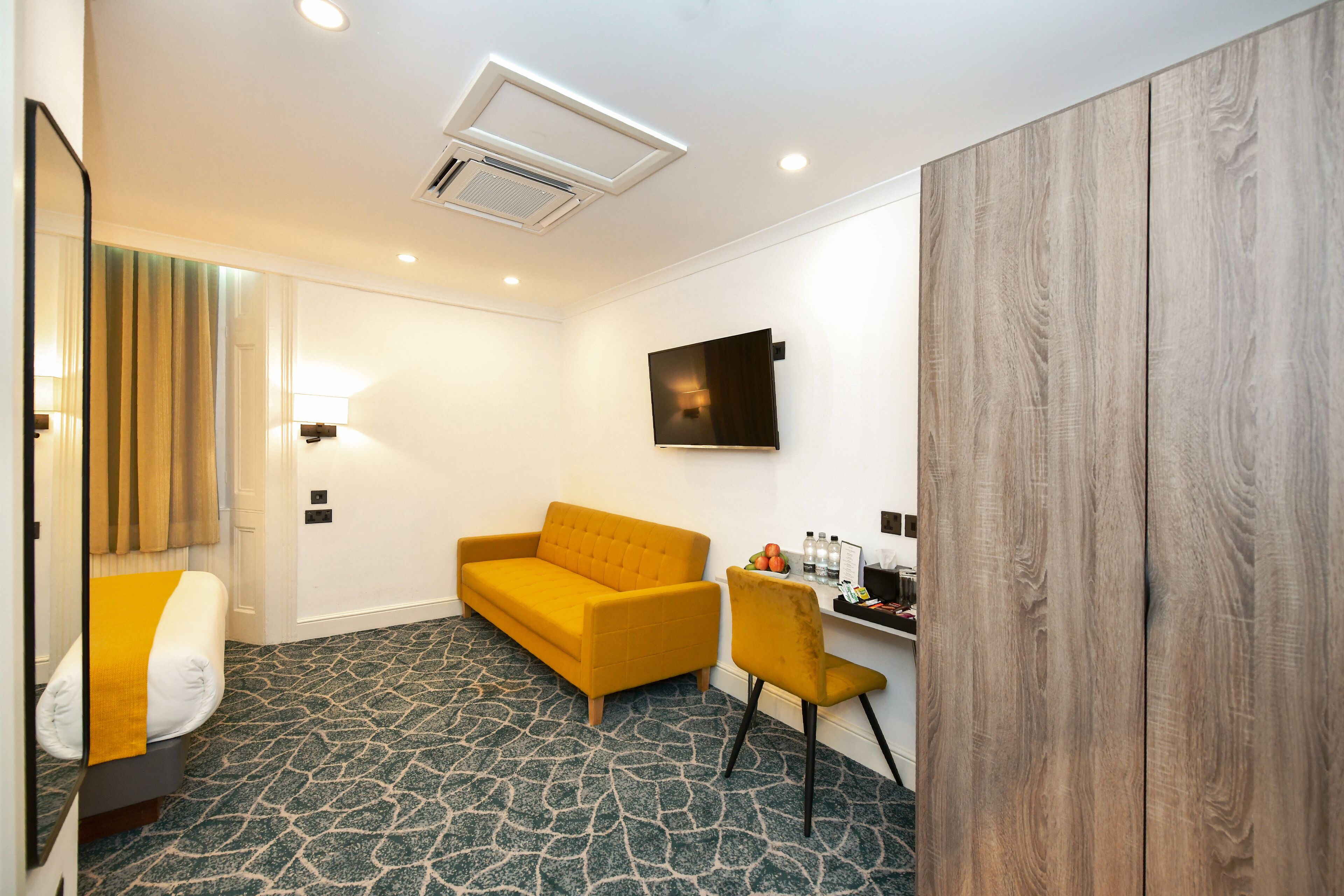 Una habitación de hotel con alfombra estampada, un sofá amarillo, un armario de madera, un escritorio con una silla y un televisor fijado a la pared.