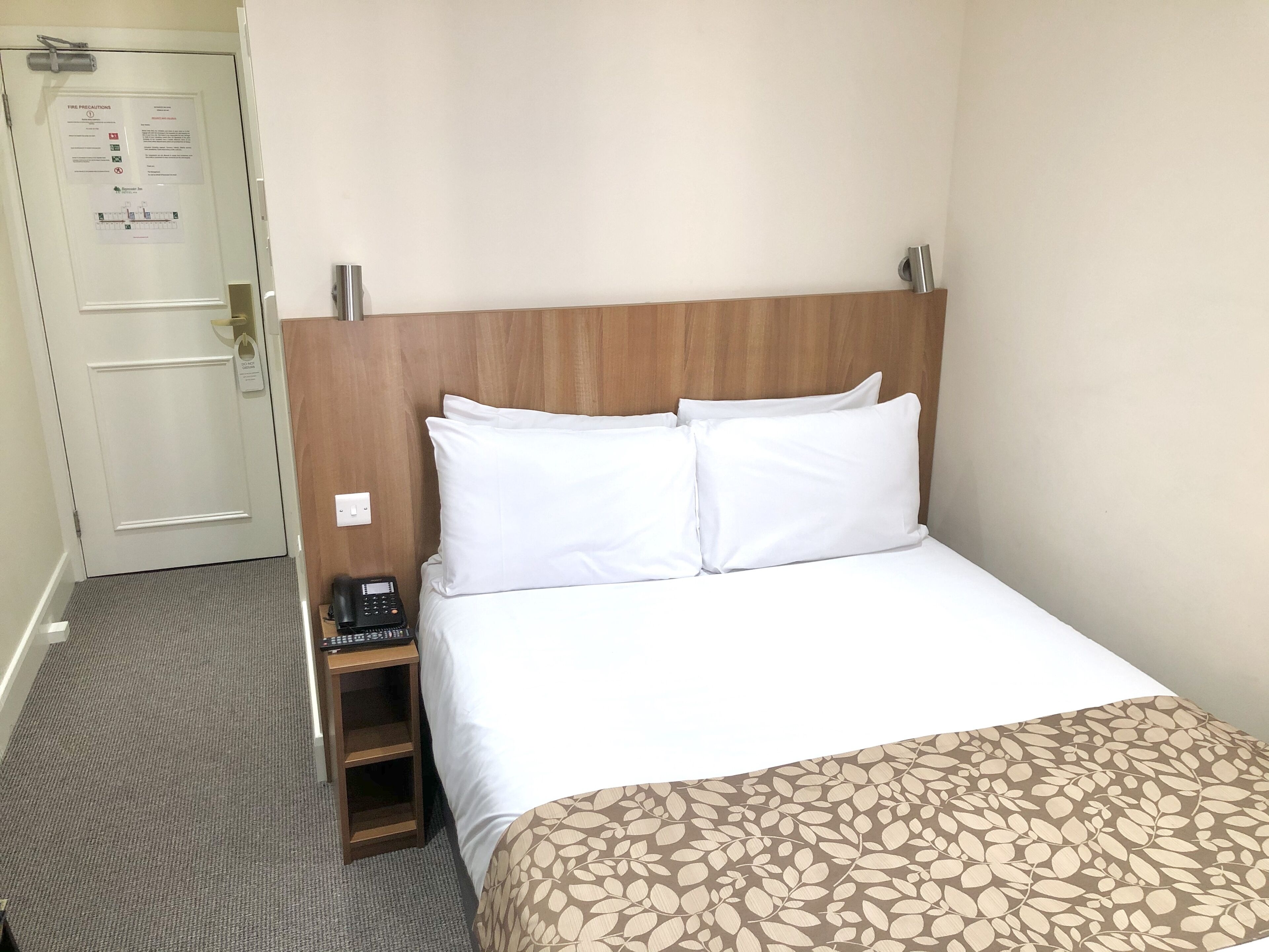 Quarto de hotel com cabeceira de madeira, roupa de cama branca, telefone ao lado da cama e chão coberto por carpete.