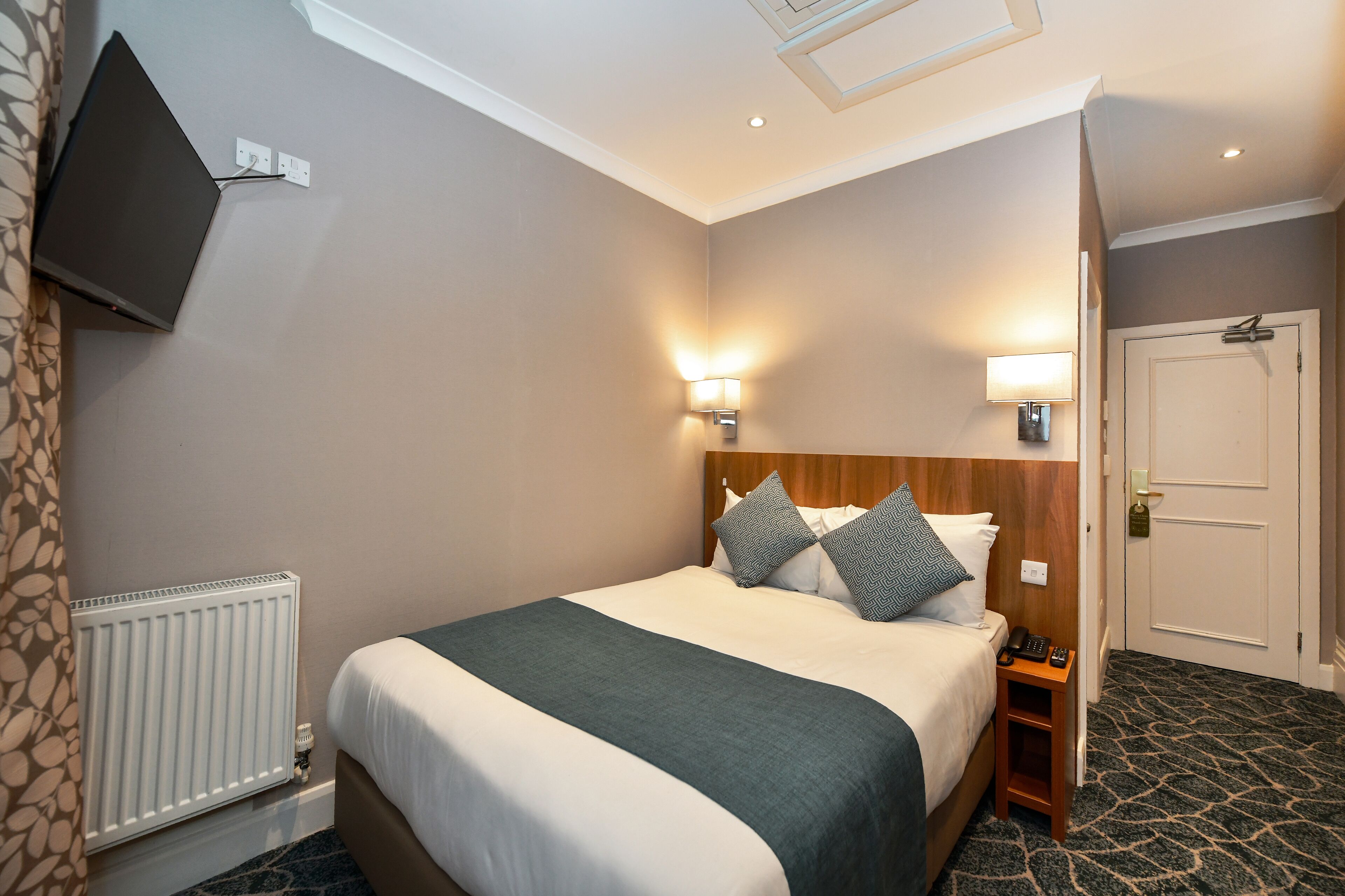 A hotel room with a bed, bedside table, lamp, and a TV mounted on the wall.