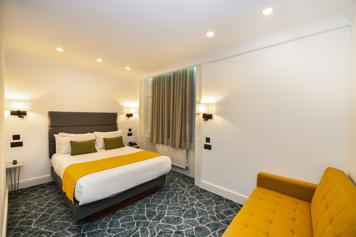 Habitació d'hotel amb llit, abacines, tauleta de nit, cadira i finestra amb cortines.