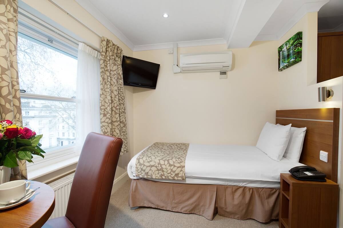 Habitació d'hotel amb llit, televisió penjada a la paret, finestra amb cortines, tauleta de nit amb telèfon i una cadira.