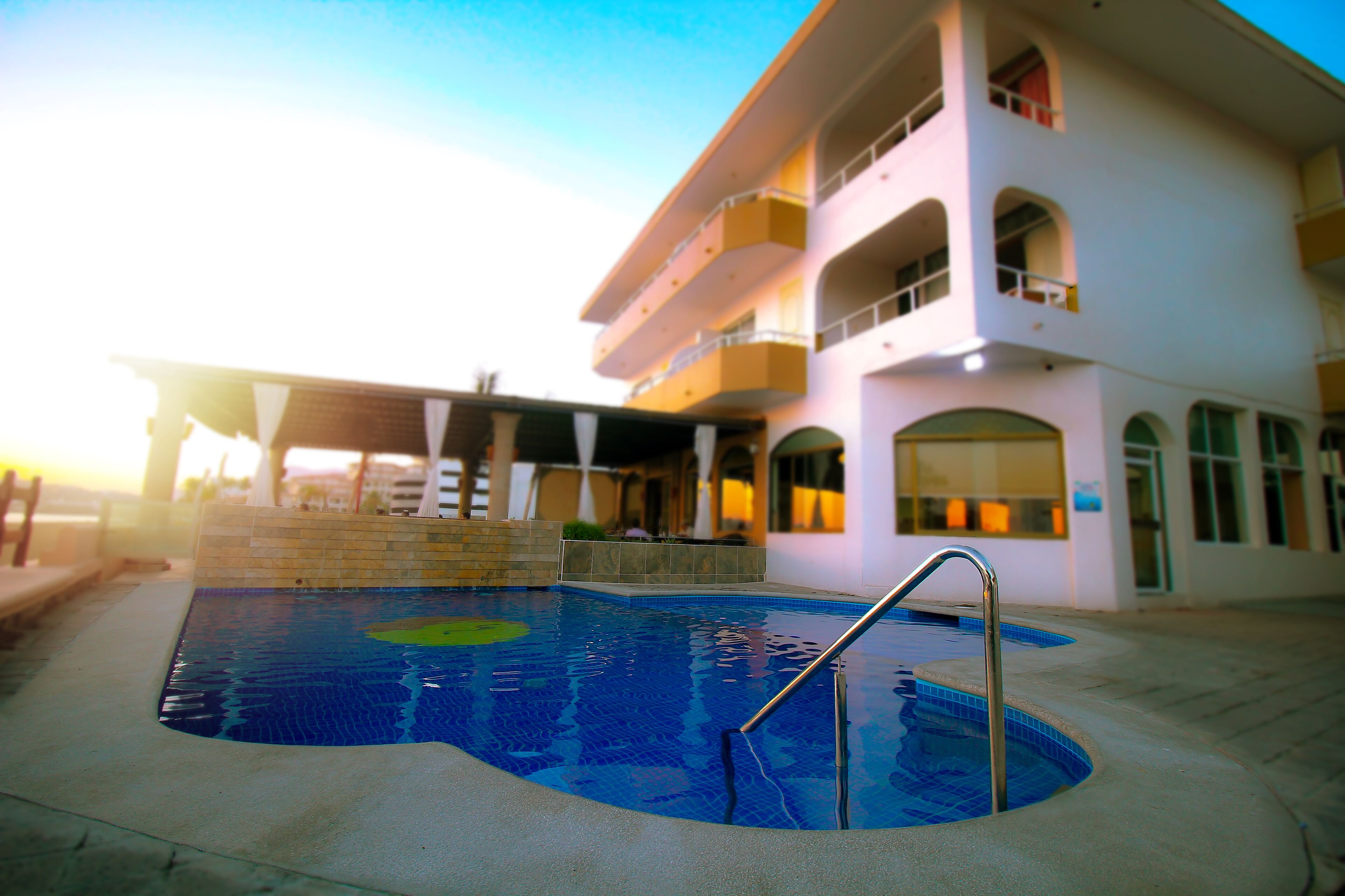 Una piscina con escalera metálica, rodeada por un solárium de hormigón, y un edificio de varios pisos con balcones al fondo.