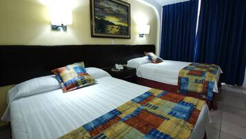 A hotel room with two beds, each with a patterned bedspread, a nightstand with a telephone, and a wall-mounted lamp.