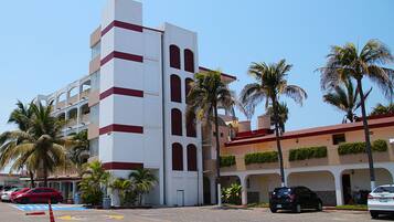 Un hotel de varios pisos con una torre central, palmeras y un estacionamiento con automóviles.