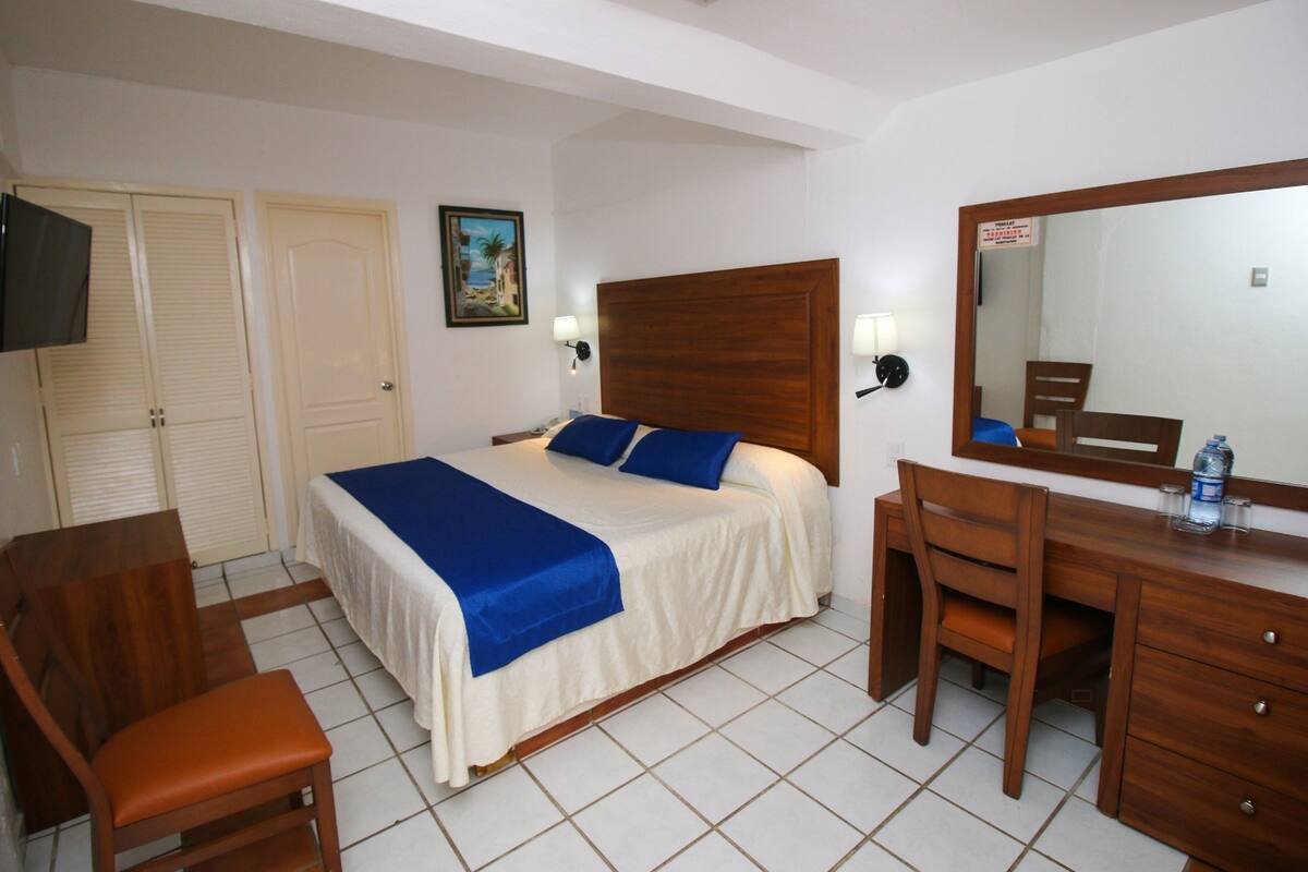 A hotel room with a bed, a wooden desk, a chair, a television, and a mirror.