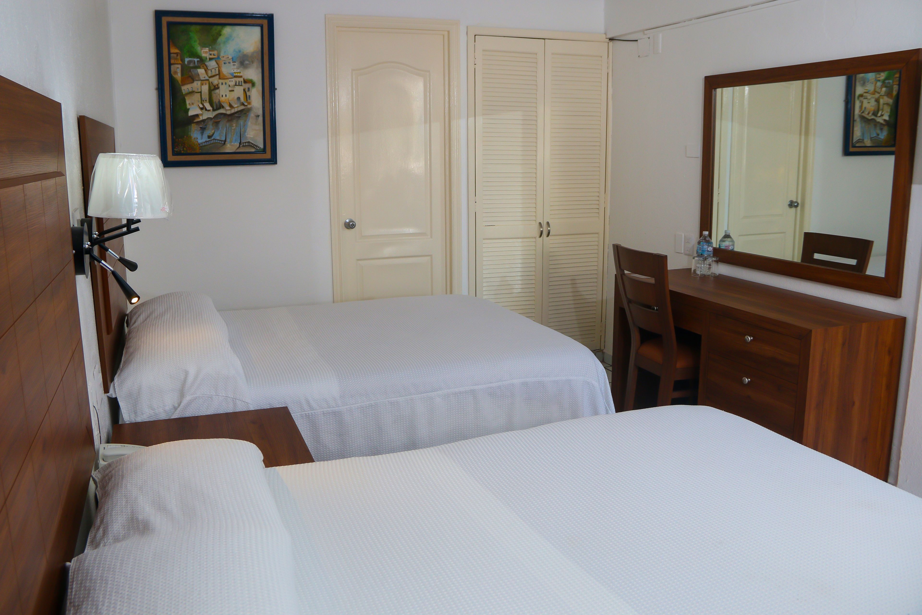 A hotel room with two beds, a desk, a chair, a dresser, a lamp, and a painting on the wall.