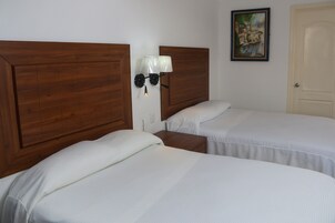 Una habitación con dos camas, cabeceros de madera, ropa de cama blanca, una mesita de noche, una lámpara de pared y un cuadro en la pared.