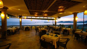 Een restaurant met tafels en stoelen, een uitzicht op zee, en hangende lampen.