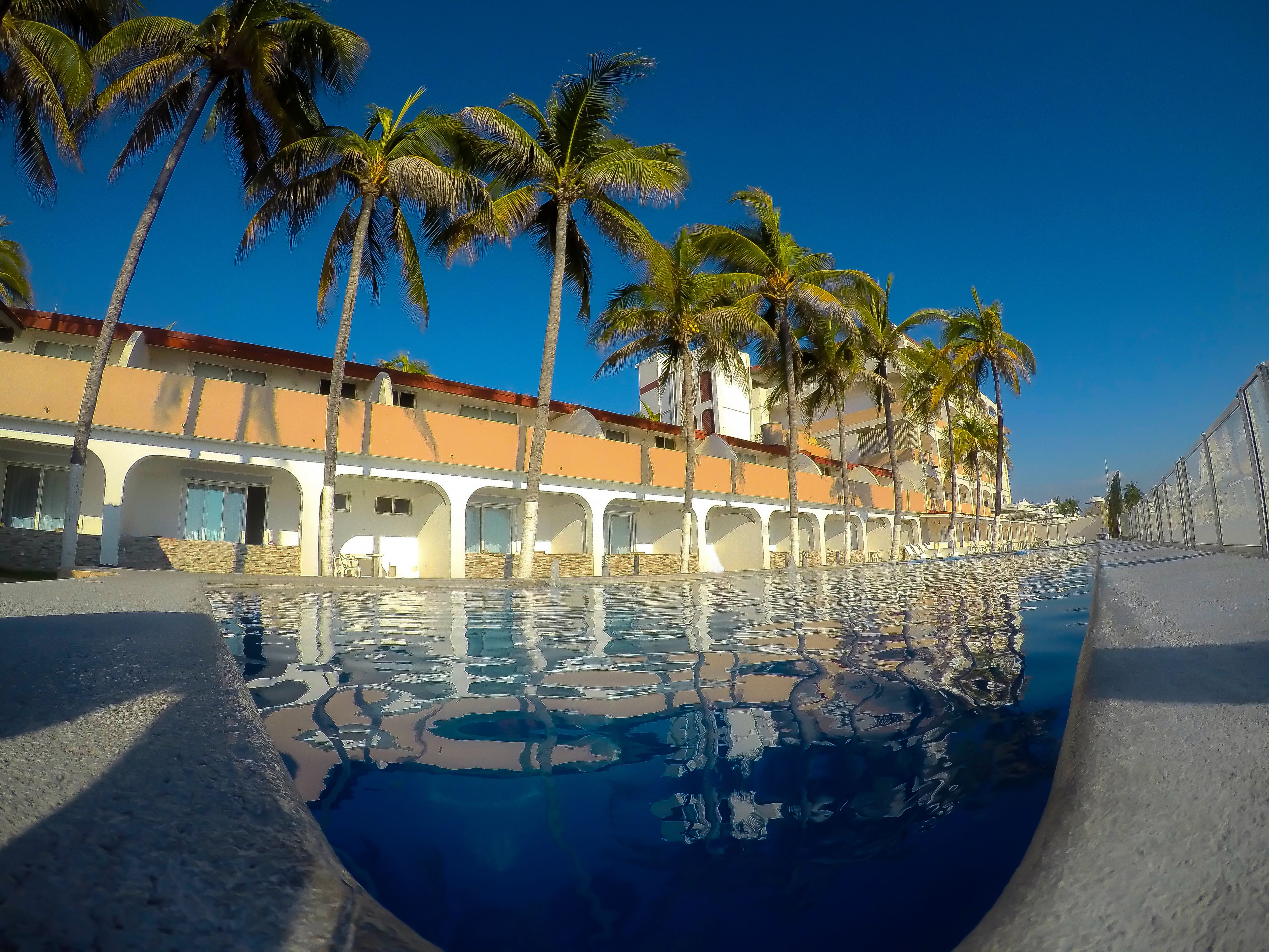 Ein Schwimmbecken mit klarem, blauem Wasser, umgeben von Palmen und einem Gebäude mit Bogenfenstern und Balkonen.