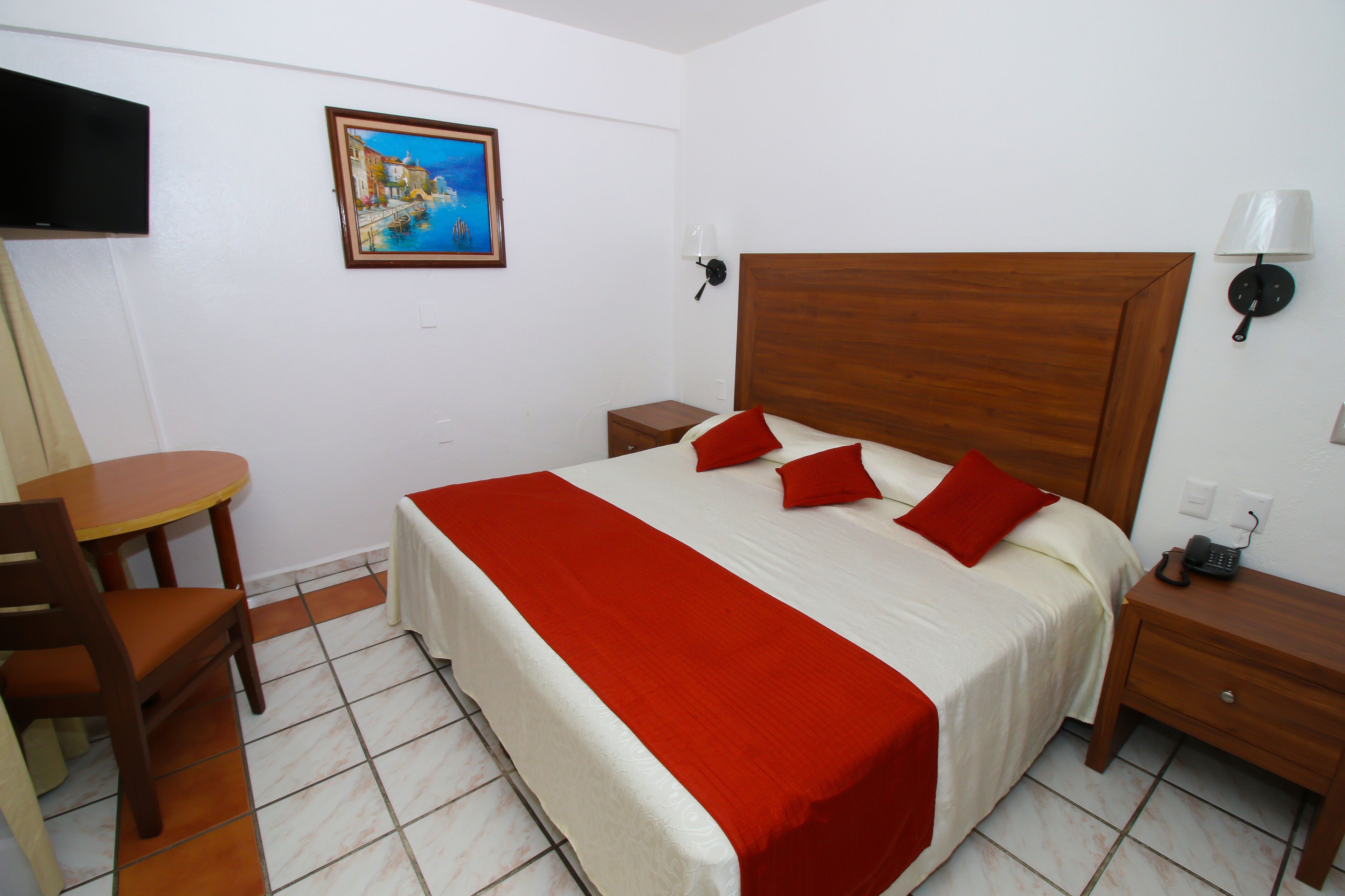 A hotel room with a bed, a bedside table, a chair, a small table, a painting, and a TV.