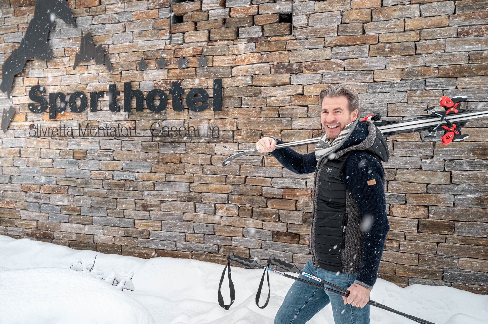 A person holding ski poles in front of a brick wall with the text "Sporthotel" and "Silvretta Montan Gaschuhi."