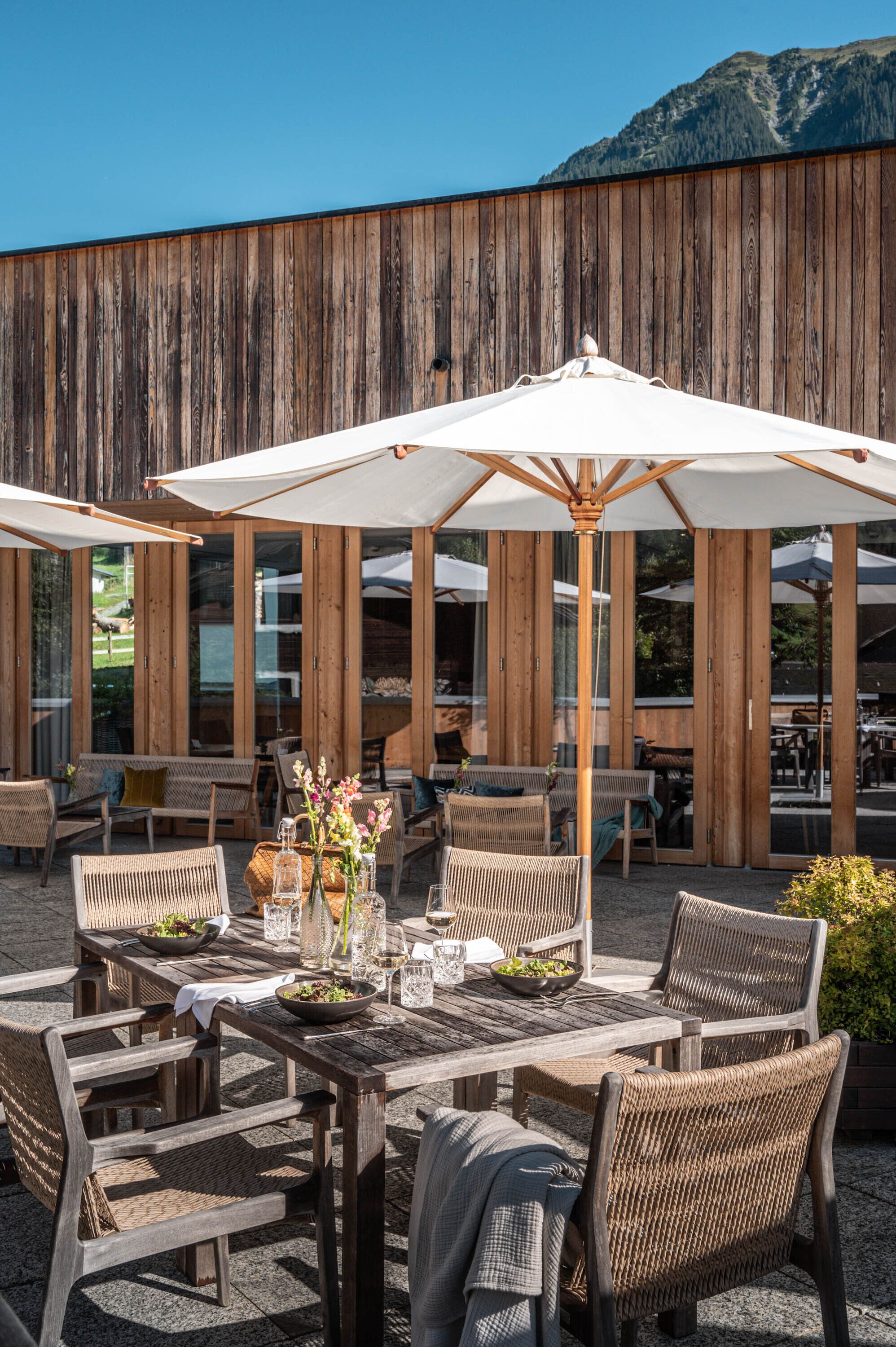 An outdoor dining area with wicker furniture and umbrellas, set against a wooden wall with large windows.